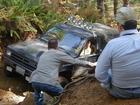 1994 Ford Explorer - A black Toyota SUV is stuck in a muddy off-road trail, with several people attempting to push it free.