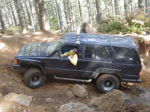 1994 Ford Explorer - A person drives a black SUV through a wooded area with rough terrain and rocks.