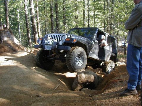 1994 Ford Explorer - A black Jeep Wrangler tackles a challenging off-road course with a person watching.