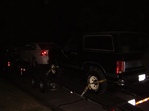 1986 Ford Bronco - Two vehicles are secured on a car hauler, being transported at night.