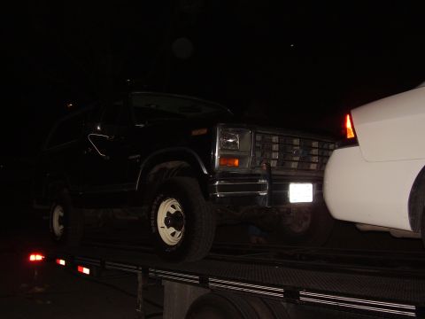 1986 Ford Bronco - A dark Ford Bronco is loaded onto a trailer at night, with a white vehicle close behind.