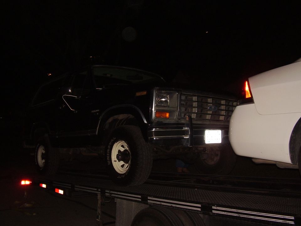 Black Ford Bronco on a trailer at night. White car is partially visible on the right
