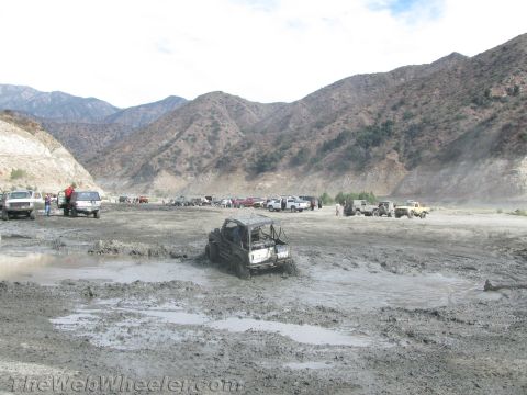 1987 Suzuki Samurai - Several off-road vehicles are stuck in a muddy area, with more vehicles parked in the background near a mountainous terrain.