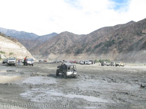 1987 Suzuki Samurai - Multiple off-road vehicles are parked and stuck in a muddy, dry riverbed with mountains in the distance.
