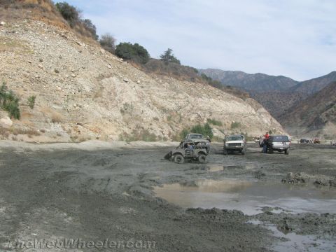 1987 Suzuki Samurai - Several off-road vehicles are seen in a muddy, dry riverbed, with steep, rocky hillsides in the background.