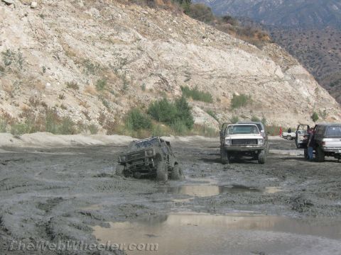 1987 Suzuki Samurai - Several off-road vehicles are stuck in deep mud near a rocky hillside.