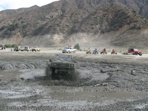 1987 Suzuki Samurai - Multiple off-road vehicles, including a 1987 Suzuki Samurai, navigate a muddy track with mountains in the background.