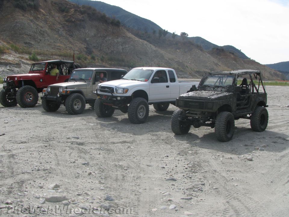Mud-covered 1987 Suzuki Samurai with roll cage parked next to a white pickup truck and two Jeeps