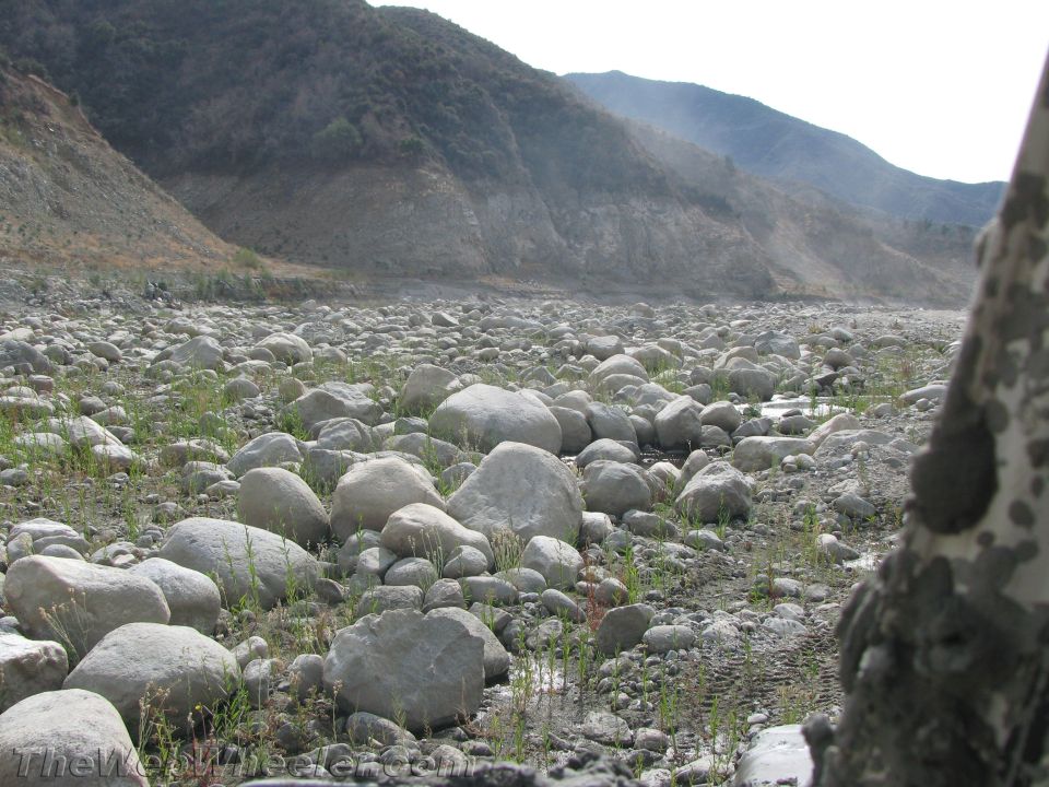 Rocky riverbed with sparse grass, leading to mountains under a hazy sky. Part of a vehicle is visible on the right