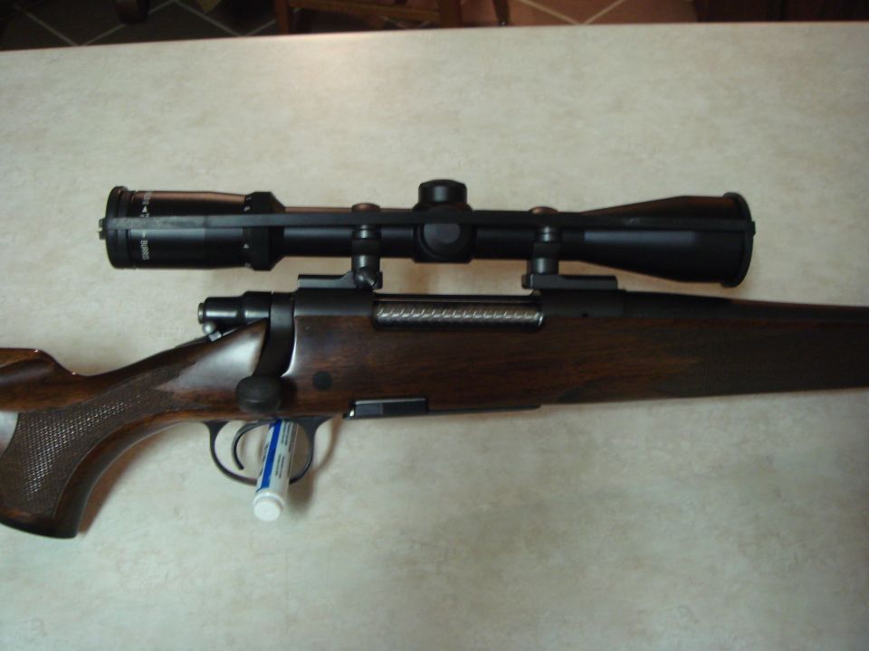 Rifle with a scope mounted on top. The rifle has a wooden stock and a metal bolt action
