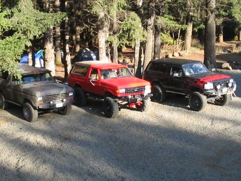 1989 Ford Bronco - Three SUVs are parked in a gravel lot surrounded by trees, with camping tents visible behind them.