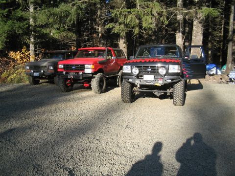 1989 Ford Bronco - Three off-road vehicles are parked on a gravel lot in a forest setting.