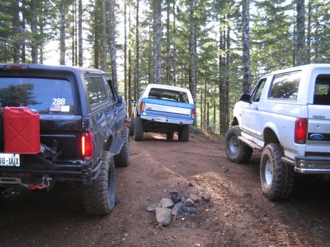 1989 Ford Bronco - Three Ford Broncos are parked on a dirt road in a forest, with a campfire pit in the foreground.
