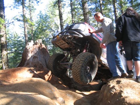 1989 Ford Bronco - An off-road vehicle navigates a challenging rocky terrain in a wooded area, with onlookers observing the ascent.