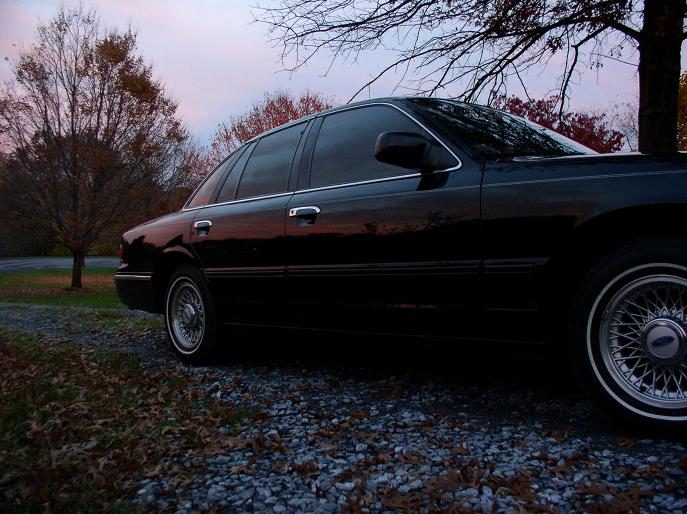 Black 1997 Ford Crown Victoria sedan, side view, parked on gravel with fall foliage