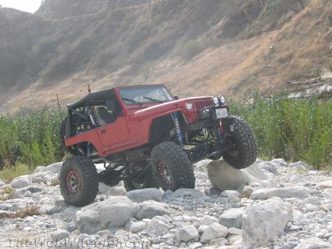 1987 Suzuki Samurai - A red Jeep Wrangler is shown off-roading over large rocks in a dry riverbed.