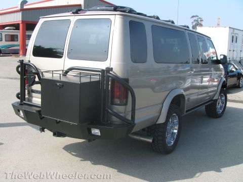 1990 Ford Bronco - Rear three-quarter view of a tan lifted Ford van with a roof rack, chrome wheels and a steel bumper holding a spare tire and box.