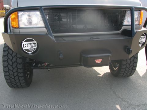 1990 Ford Bronco - Close-up of a tan van's black steel front bumper fitted with round Hella driving lights, a Warn winch and a grille brush guard.