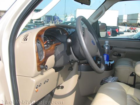 1990 Ford Bronco - Interior of a Ford van showing the tan leather seats, woodgrain dash trim, and a Ford-emblem steering wheel with a water bottle in the door.