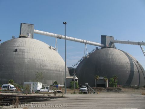 1990 Ford Bronco - Two enormous gray dome storage buildings connected by elevated conveyor tubes, with trucks parked at an industrial port facility.