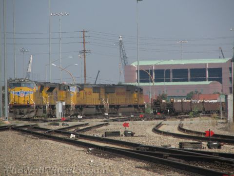 1990 Ford Bronco - Three yellow Union Pacific diesel locomotives sit coupled in a rail yard with signal lights, switch tracks, and port cranes in the distance.