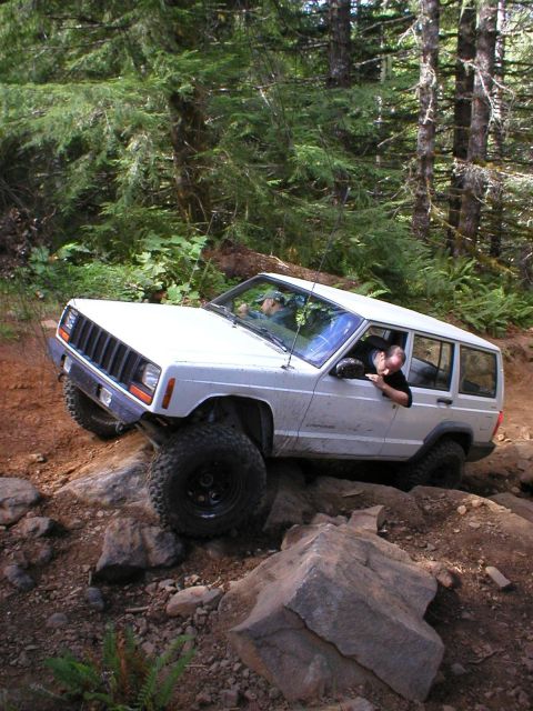 2005 Jeep Wrangler - A white Jeep Cherokee XJ is shown off-roading on a rocky trail surrounded by trees.