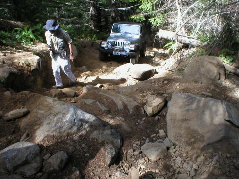 2005 Jeep Wrangler - A Jeep Wrangler tackles a challenging off-road path with large rocks and trees surrounding it.
