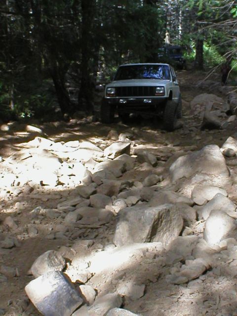 2005 Jeep Wrangler - White Jeep Cherokee XJ navigates a challenging, rocky trail in a forest setting with another vehicle following.