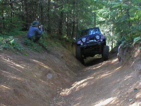 2005 Jeep Wrangler - A blue Jeep Wrangler navigates a challenging dirt path in a wooded area, observed by a photographer.