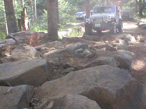 2005 Jeep Wrangler - Multiple Jeeps traverse a challenging, boulder-strewn path in a wooded area.