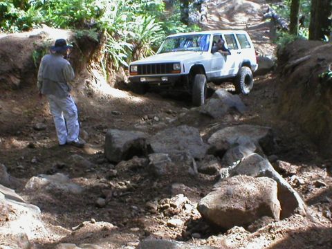 2005 Jeep Wrangler - A white Jeep Cherokee XJ navigates a challenging, rocky off-road path with a person watching.