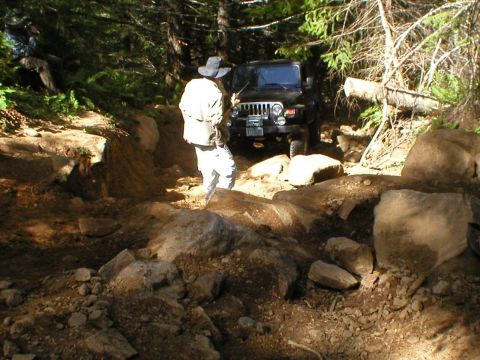 2005 Jeep Wrangler - A black Jeep Wrangler tackles a challenging rocky path in a wooded area, with a person observing the off-road adventure.
