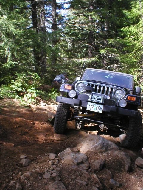 2005 Jeep Wrangler - A black 2005 Jeep Wrangler tackles a challenging rocky path through a dense forest, with another vehicle visible in the background.