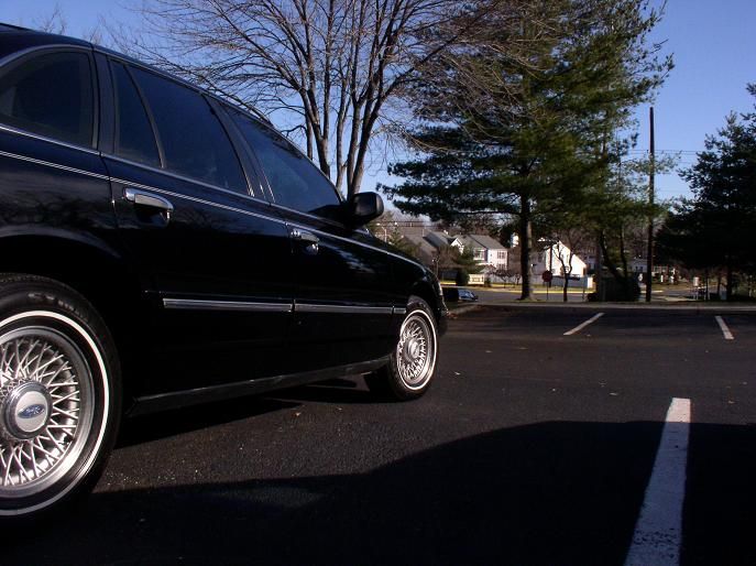 Side view of a black 1997 Ford Crown Victoria parked in a lot with trees and houses in the background