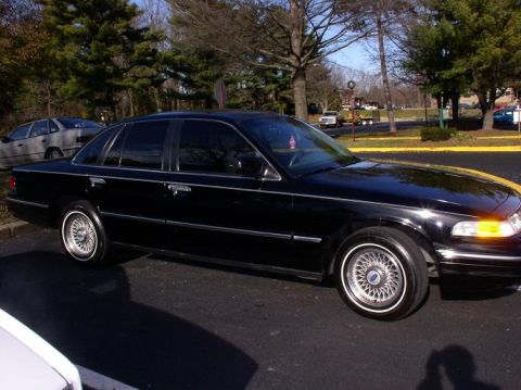 1997 Ford Crown Victoria - A black 1997 Ford Crown Victoria is parked on an asphalt surface with trees in the background.