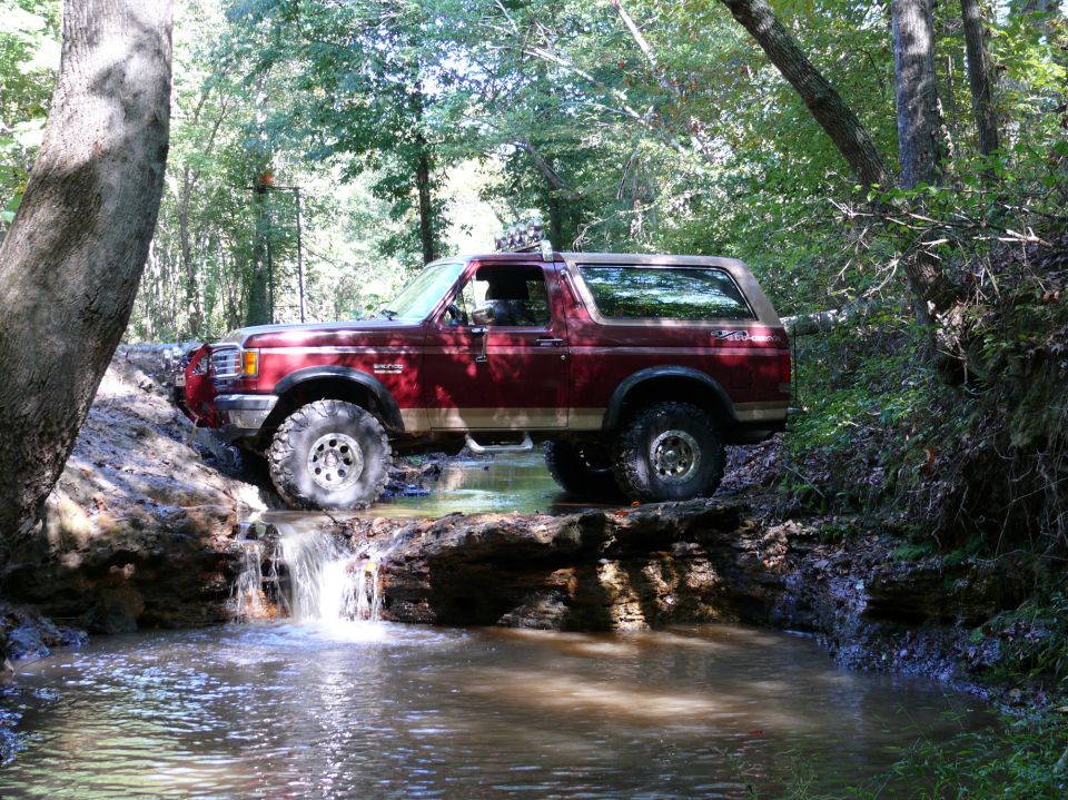Red Ford Bronco with off-road tires crossing a stream on a log in a wooded area
