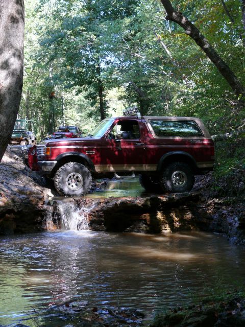 1988 Ford Bronco - A red Ford Bronco navigates a water obstacle on a wooded trail, with another vehicle in the background.