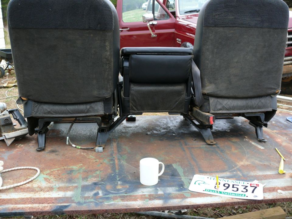 Two dark fabric car seats with a center console, on a rusty surface, with a red vehicle in the background