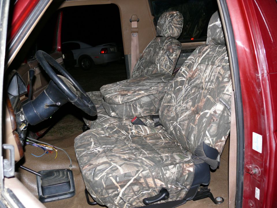 Interior of a red vehicle with camouflage seat covers, steering wheel, and exposed wires. A white car is visible outside