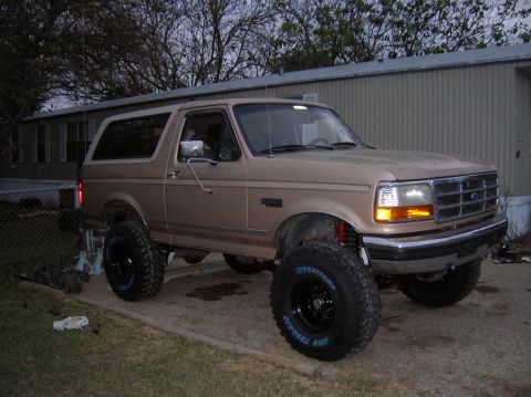 1996 Ford Bronco - Passenger-side front three-quarter view of a freshly lifted tan Ford Bronco parked at dusk in front of a metal building, with 'BRONCO' fend…