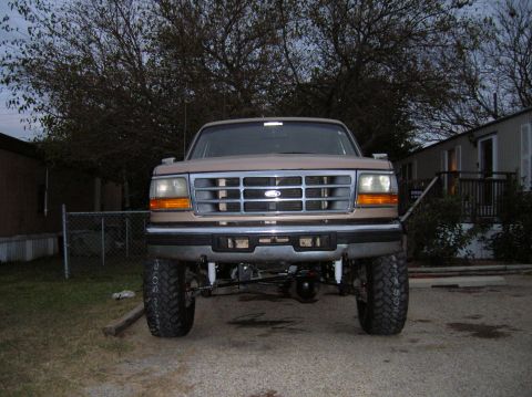 1996 Ford Bronco - Low head-on view of a tan lifted Ford SUV at dusk in a gravel driveway, showing the chrome slotted grille with blue oval emblem, chrome bum…