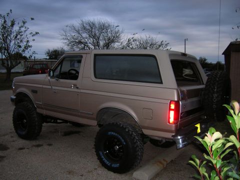 1996 Ford Bronco - Driver-side rear three-quarter view of a lifted tan Ford two-door SUV with its removable hardtop in place, parked on gravel under an overca…