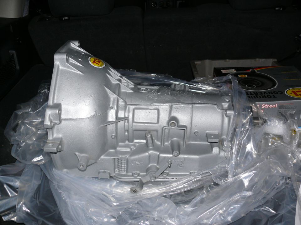 Silver transmission part in plastic wrap, with a yellow sticker and a box labeled 'TORQUE CONVERTER' in the background