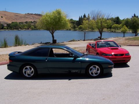 1996 Ford Probe - A green coupe is parked next to a red convertible with a lake and trees in the background.