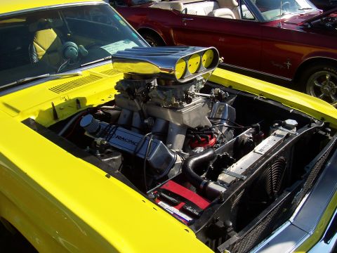 1973 Ford Mustang - The engine bay of a yellow 1973 Ford Mustang features a prominent chrome scoop and dual carburetors, with another classic car visible in th…