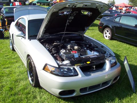 1973 Ford Mustang - A silver Ford Mustang Cobra is displayed with its hood open, revealing the engine and "COBRA" lettering on the hood's interior.