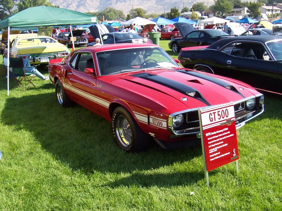 Red 1970 Shelby Mustang GT500 with black stripes and GT500 side decals, parked on grass at a car show
