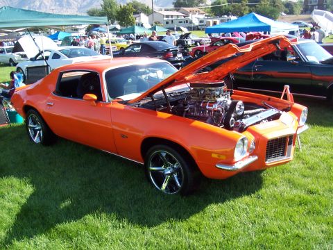 1973 Ford Mustang - This orange 1970 Chevrolet Camaro features a prominent supercharged engine under its open hood, displayed at an outdoor car show.