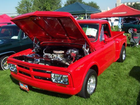 1973 Ford Mustang - A bright red pickup truck is displayed with its hood open, revealing the engine bay. The vehicle is on display at an outdoor event.