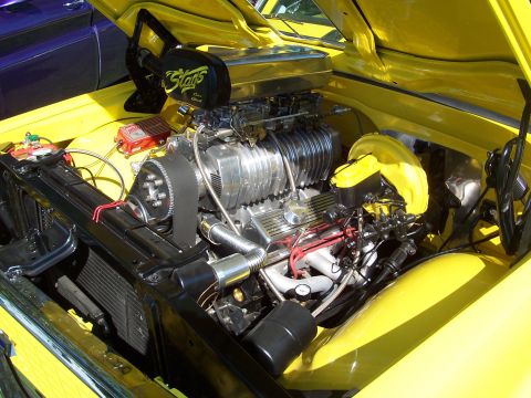 1973 Ford Mustang - The engine bay of a yellow vehicle displays a prominent supercharger and dual carburetors, with another vehicle visible in the background.